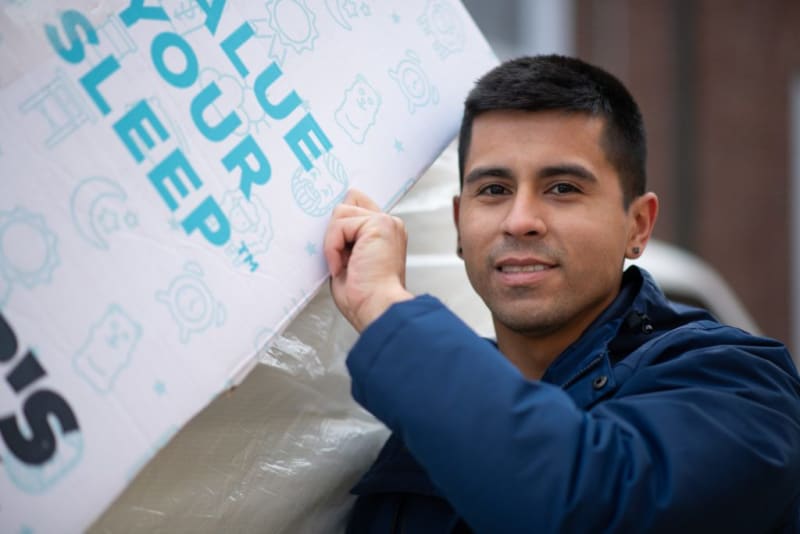 Person holding a white mattress box with blue text "LOVE YOUR SLEEP" and small sleep-themed icons, wearing a blue jacket.