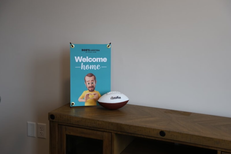 Blue sign with white text "Welcome home" and a cartoon character wearing a yellow shirt, placed on a wooden surface next to a white and brown football.