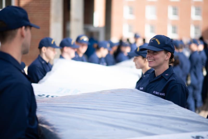 U.S. Coast Guard personnel in navy blue uniforms and caps working together to carry a large covered object outdoors.