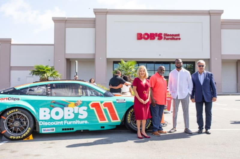 Four people stand beside a turquoise and orange race car with "BOB'S Discount Furniture" branding and number 11, in front of a BOB'S Discount Furniture store.