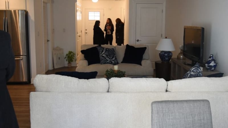 Modern living room with beige fabric sofas, navy and patterned throw pillows, hardwood floors, stainless steel refrigerator, and three people conversing near the entrance.