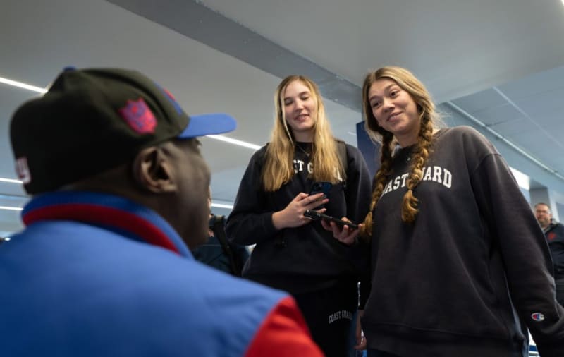 Two people wearing black Coast Guard sweatshirts, one with braided hair, holding smartphones indoors under fluorescent lighting.