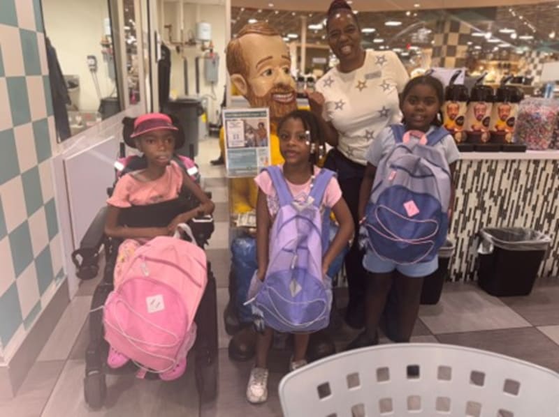 Three children with backpacks standing indoors near a retail display; one child in a wheelchair with a pink backpack, another with a purple backpack, and a third with a blue backpack.