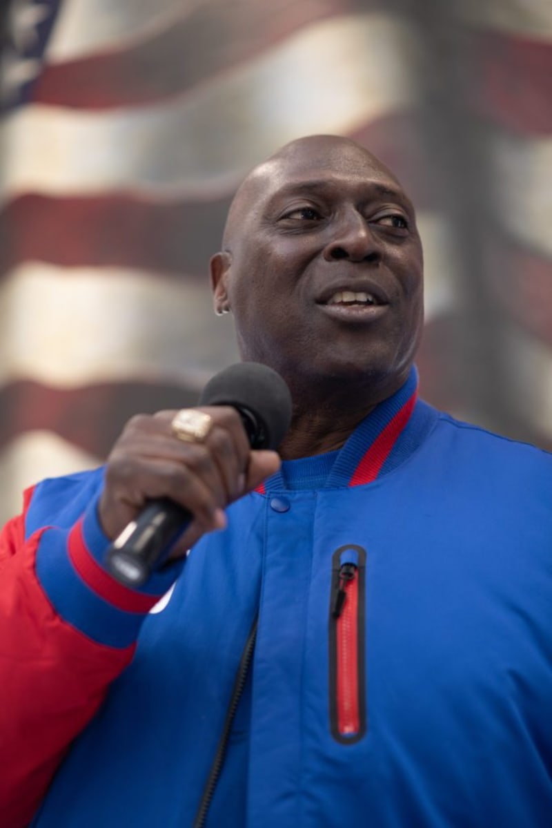 Person wearing a blue and red jacket holding a microphone, with a blurred American flag in the background, suggesting a speech or public event.