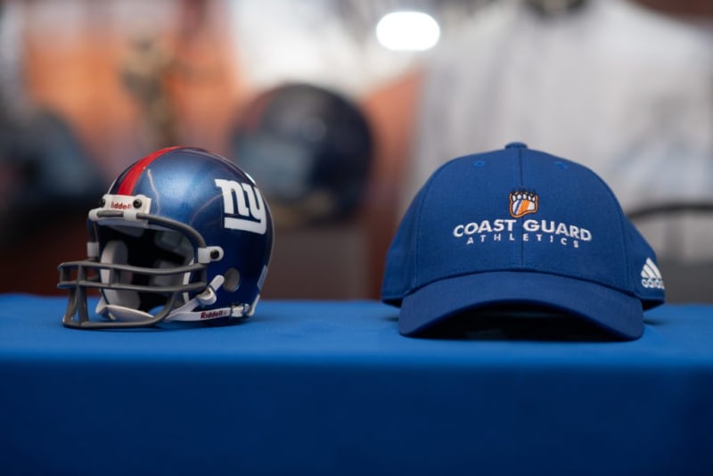 Blue Riddell football helmet with New York Giants logo and a blue Adidas Coast Guard Athletics cap with orange and white logo, both on a blue table.