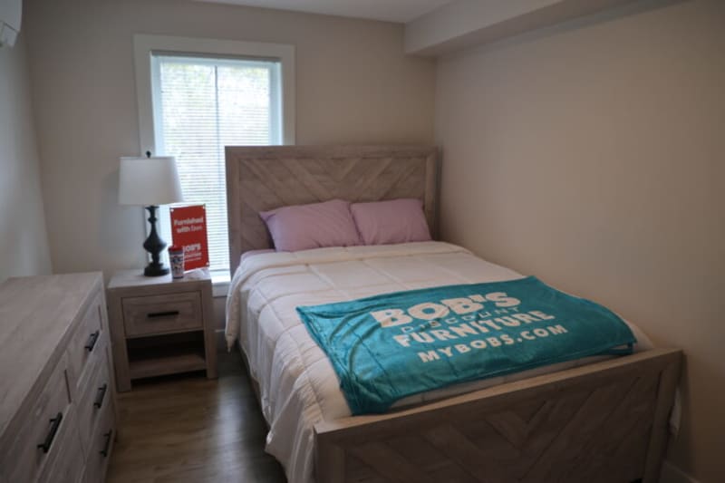 Bedroom setup featuring a wooden bed with a chevron pattern headboard and footboard, white bedding, purple pillows, and a teal blanket with "BOB'S DISCOUNT FURNITURE MYBOBS.COM" text. Includes a wooden nightstand with a black lamp and a dresser, showcasing modern, rustic furniture style.