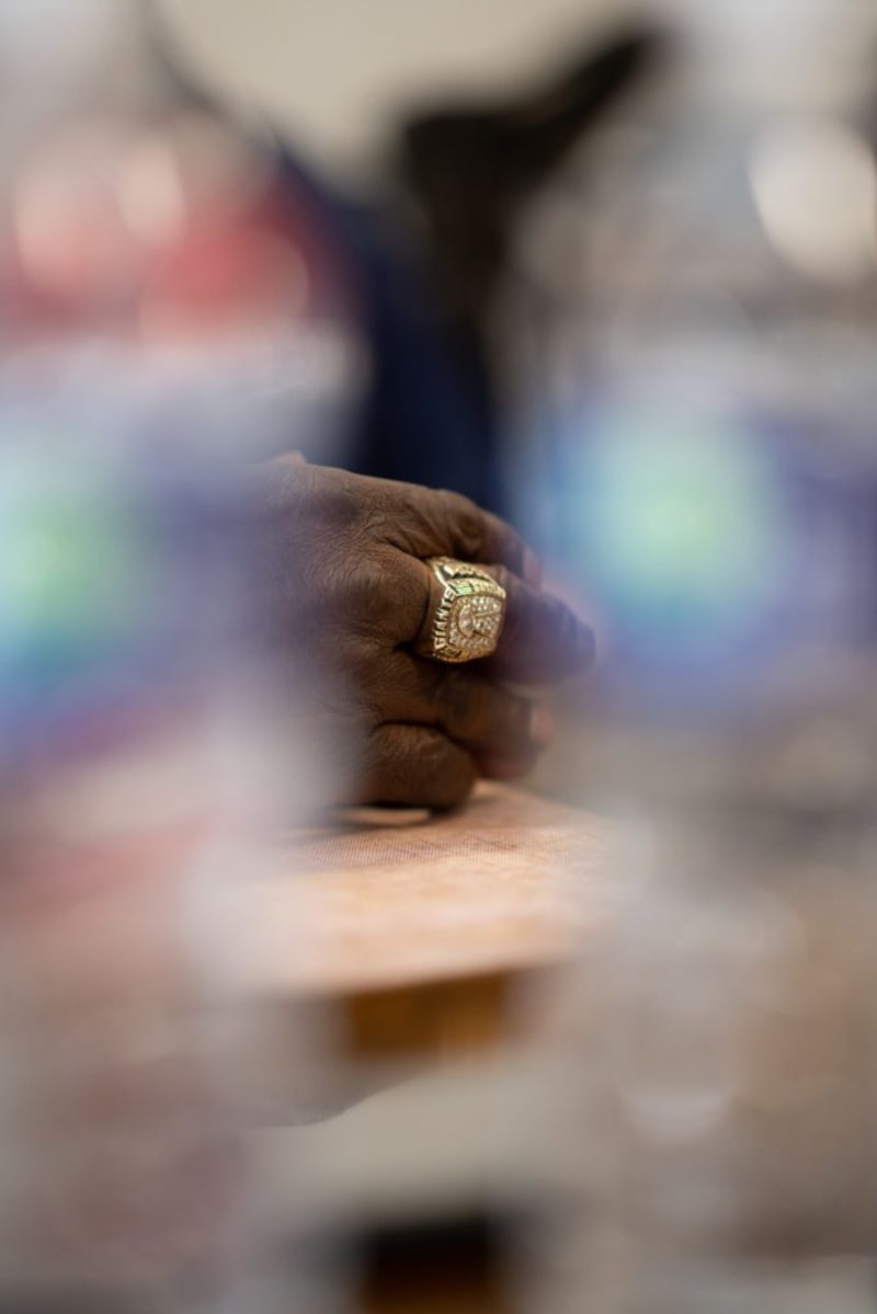 Dark-skinned hand wearing a large, gold championship ring with intricate designs, resting on a wooden surface, framed by blurred water bottles.
