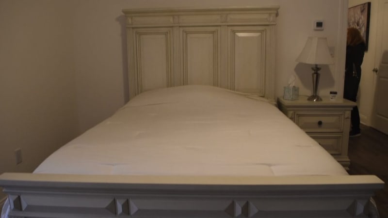 Queen-sized bed with cream-colored wooden headboard and footboard, white comforter, matching nightstand with lamp and tissue box in a bedroom.