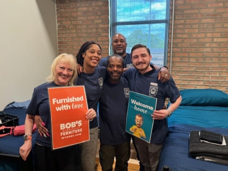 A group of five people wearing matching navy blue shirts stand in a furnished room with a bed and window. Two hold signs: one orange with white text reading "Furnished with love BOB'S DISCOUNT FURNITURE MYBOBS.COM," and the other teal with white text reading "Welcome home." The setting suggests a home furnishing or charity event promoting Bob's Discount Furniture.
