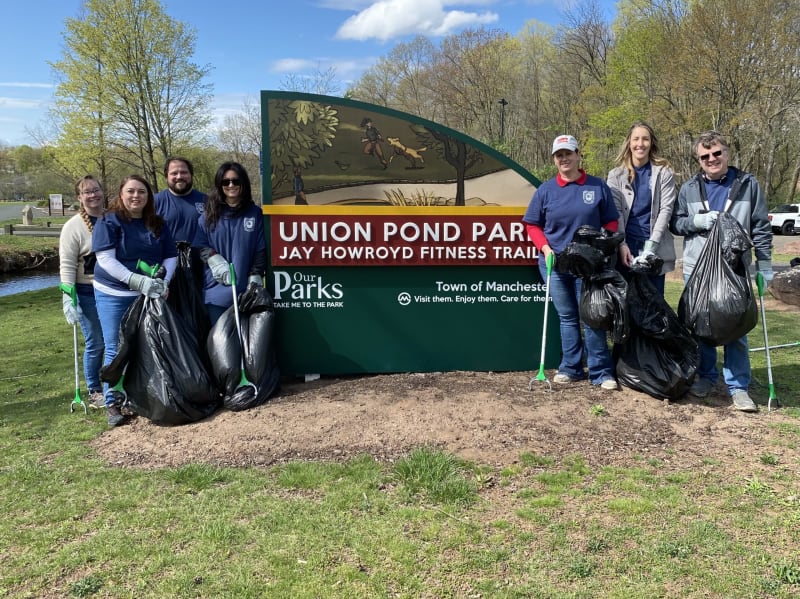 Group of people holding black trash bags and litter pickers standing in front of a green sign reading "Union Pond Park Jay Howroyd Fitness Trail Our Parks Town of Manchester Visit them. Enjoy them. Care for them."