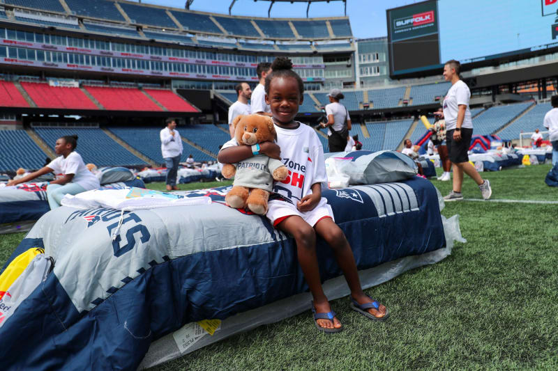 Child sitting on a New England Patriots-themed sleeping bag on a football field, hugging a teddy bear wearing a Patriots sweatshirt, with other children and adults in the background.