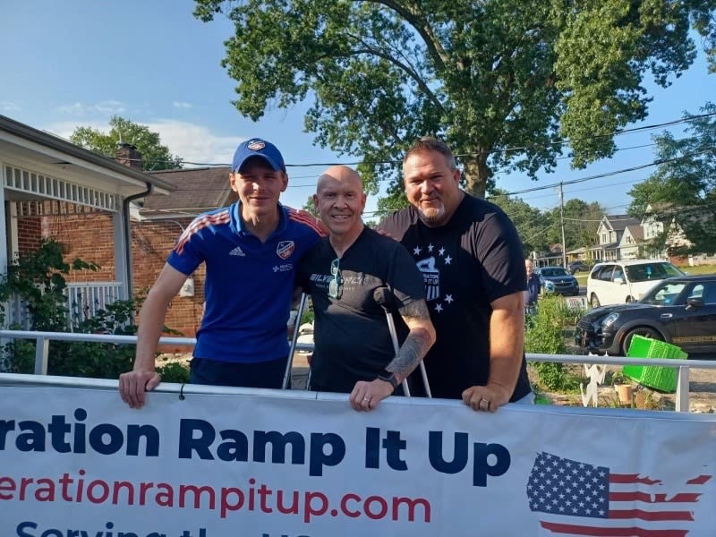 Three men standing behind a banner that reads "Operation Ramp It Up" and "operationrampitup.com" with an American flag graphic, outdoors on a sunny day.