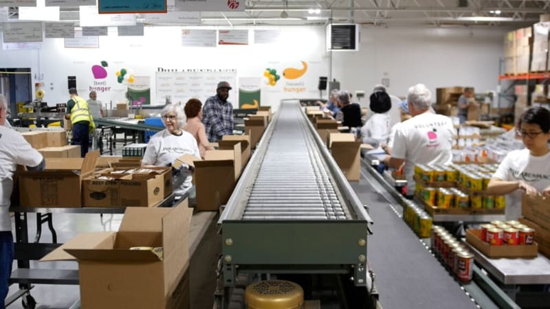 Volunteers packing food items into boxes on a conveyor belt at a Philadelphia-based hunger relief warehouse with "beet hunger" and "peach hunger" logos.