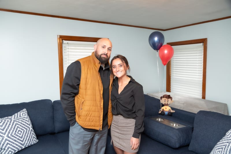 A man wearing a mustard quilted vest and gray pants stands next to a woman in a black blouse and plaid skirt in a living room with blue couches, patterned pillows, and two balloons (red and blue) attached to a plush toy.
