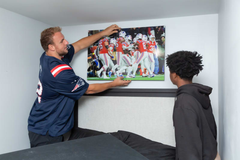 A person wearing a navy blue New England Patriots jersey hangs a framed football team photo above a bed with a beige headboard, while another person watches.