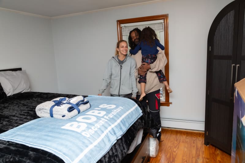 A bedroom with a black plush bedspread and a light blue fleece blanket labeled "BOB'S Discount Furniture," featuring white pillows and folded towels on the bed, with three people standing near a window and a dark wooden wardrobe on the right.