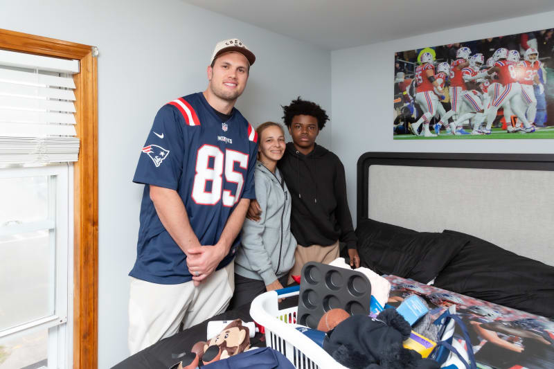 Three people standing in a bedroom with a New England Patriots jersey, casual clothing, and a baseball cap. The bed has a sports-themed comforter and a laundry basket with household items. A football game poster is on the wall.