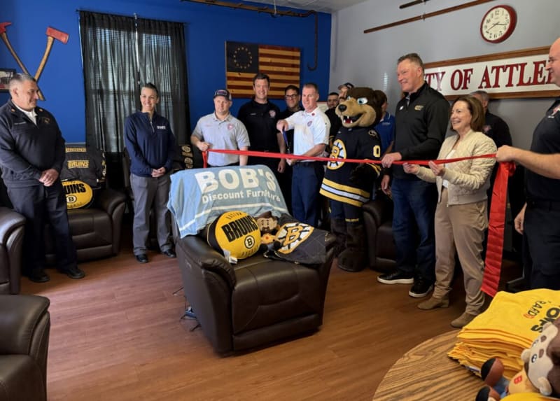 Group of people, including a mascot in a Boston Bruins hockey jersey, holding a red ribbon for a ribbon-cutting event in a room with Bruins-themed furniture and decor. The room features a chair covered with a blue towel reading "BOB'S Discount Furniture," Bruins pillows, and a sign partially reading "CITY OF ATTLE."