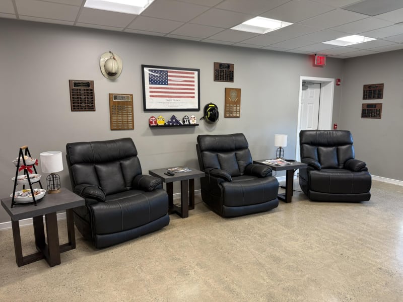 Waiting area with three black leather recliner chairs and dark wood side tables, decorated with firefighter helmets, plaques, and an American flag wall art.