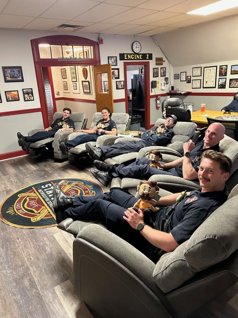 Four firefighters relaxing on gray recliner chairs in a fire station lounge with "ENGINE 2" sign and fire department emblem rug on wood flooring.