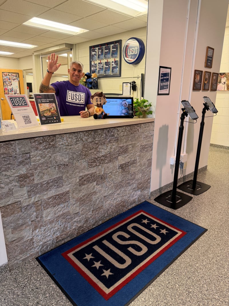 USO service desk with a staff member in a purple USO shirt waving behind a stone-textured counter, USO logo mat on floor, and informational signs.