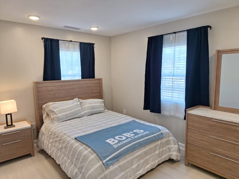 Bedroom setup with wooden bed frame, striped bedding, and a blue throw blanket reading "BOB'S Discount Furniture." Includes matching wooden nightstand with lamp and dresser with mirror. Navy blue curtains with white sheers on windows.