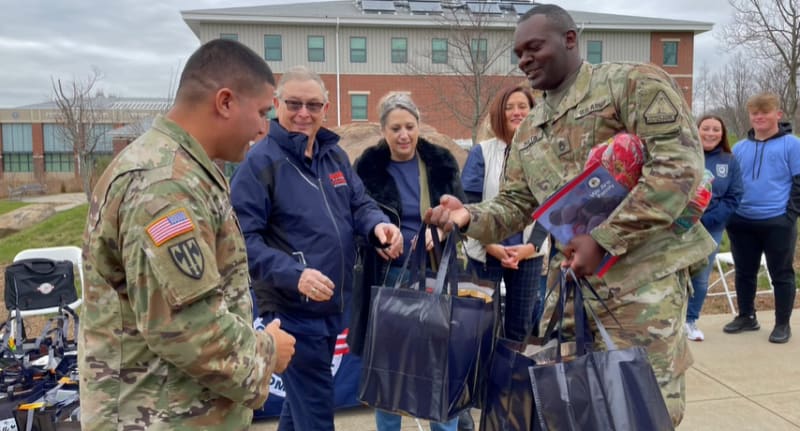 Two U.S. Army soldiers in camouflage uniforms hand out black reusable bags to two women outdoors near a brick building, with others watching.