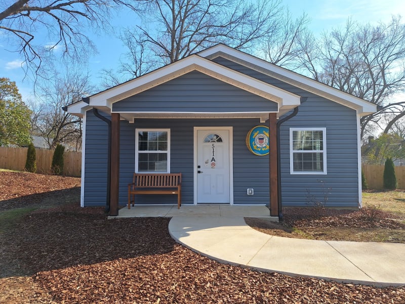 Small blue house with white trim and a white door labeled "511A," featuring a wooden bench on the front porch and a curved concrete walkway.