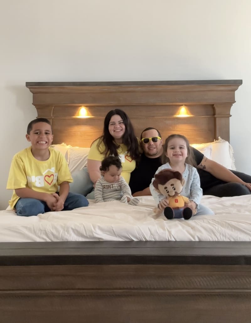 A family of five sitting on a large wooden bed with white bedding, smiling and holding a plush toy, in a well-lit bedroom.