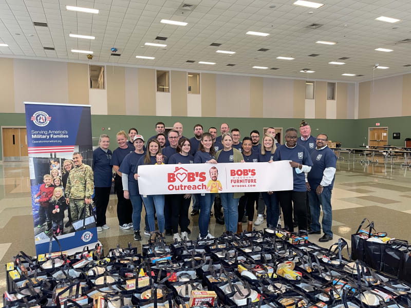 Group of volunteers standing indoors behind a banner reading "BOB'S Outreach | BOB'S Discount Furniture MYBOBS.COM" with bags of food and supplies in front, supporting military families.