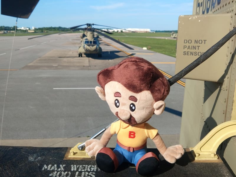 Plush doll with brown hair, mustache, yellow shirt with red "B," and blue pants sitting inside a military helicopter with "DO NOT PAINT SENSOR" text visible.