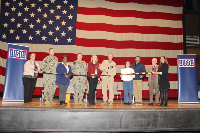 A group of military personnel and civilians stand on stage in front of a large American flag, holding a ceremonial hockey stick for a USO event.