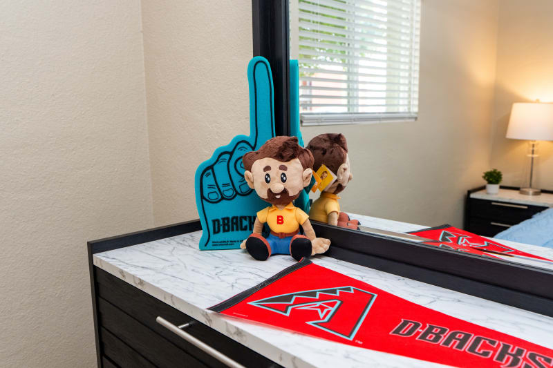 Plush toy with brown hair and beard wearing a yellow shirt with red "B" and blue pants, sitting next to a blue foam finger with "DBACKS" text and a red Arizona Diamondbacks pennant on a marble surface in front of a mirror.