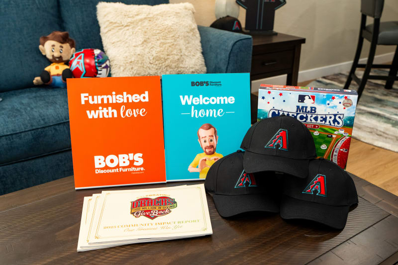 Marketing display on a wooden table featuring two Bob's Discount Furniture signs with texts "Furnished with love" and "Welcome home," three black Arizona Diamondbacks caps, MLB Checkers game, and D-Backs 2025 Community Impact Report.