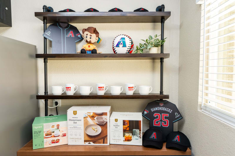 Shelf display with Arizona Diamondbacks baseball merchandise including black jerseys with red and teal accents, caps, a plush doll, a baseball, and white mugs with "BOB'S FURNITURE" logo, alongside boxed kitchenware sets labeled "Threshold" and "Figmint Tritan Food Storage Set."