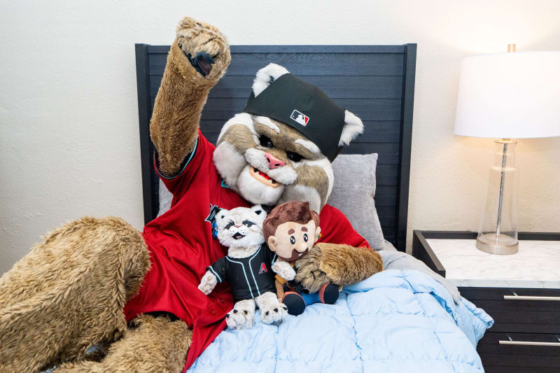A person in a furry mascot costume wearing a red jersey and black cap sits on a bed holding two plush toys, one resembling the mascot.