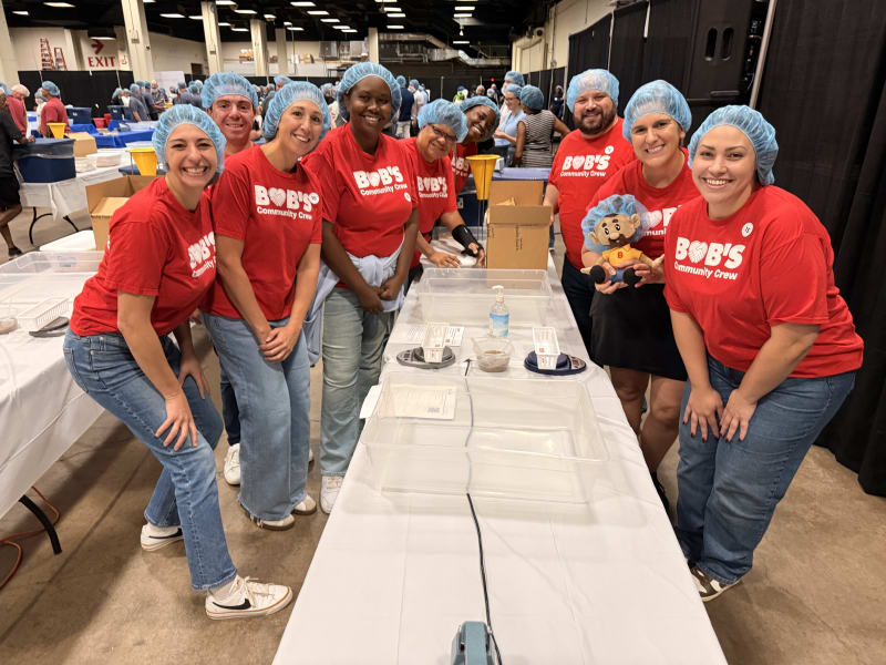Group of volunteers wearing blue hairnets and red "BOB'S Community Crew" t-shirts standing around a table with plastic trays and food packaging, likely participating in a food packing or community service event.