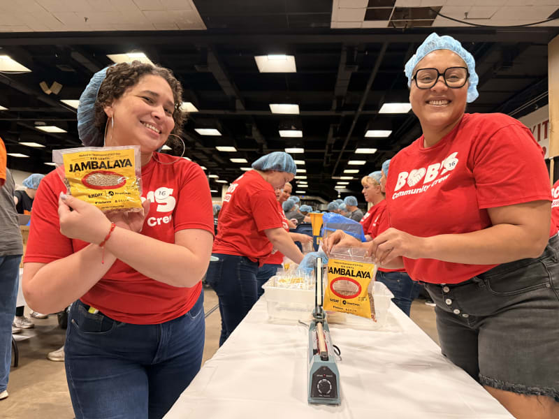Two women wearing red "Bobe Community Crew" shirts and blue hairnets smile while holding and packaging yellow bags of red lentil jambalaya in a food packing event.