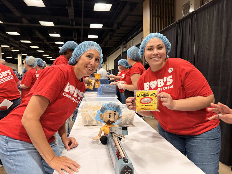Two women wearing blue hairnets and red "BOB'S Community Crew" t-shirts smile while packaging yellow "JAMBALAYA" seasoning packets at a volunteer event.