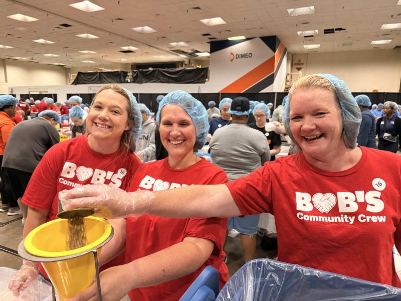 Three women wearing red "BOB'S Community Crew" t-shirts and blue hairnets are smiling while pouring food into a yellow funnel for packaging at a community food packing event.