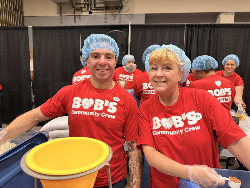 Two volunteers wearing red "BOB'S Community Crew" t-shirts and blue hairnets, working at a food packaging event with gloves and containers.