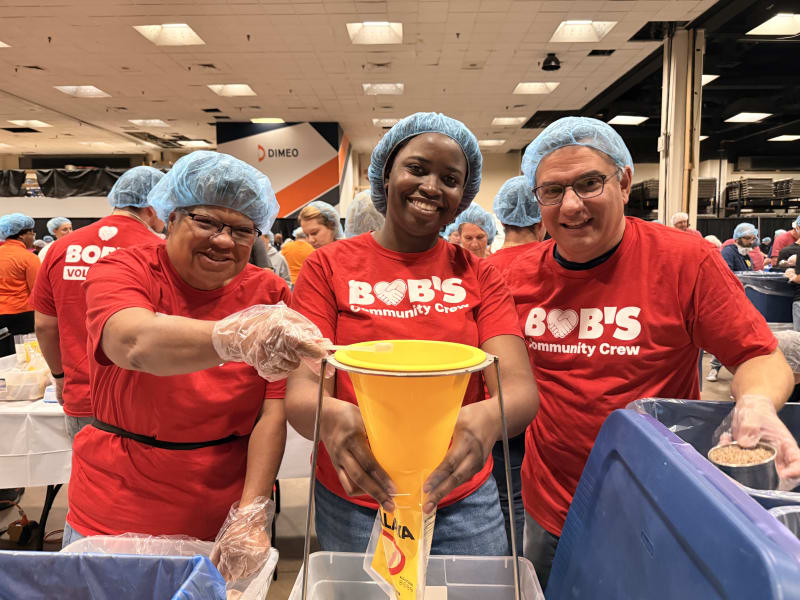 Three volunteers wearing red "Bob's Community Crew" t-shirts and blue hairnets pack food using a yellow funnel in a large indoor event space.