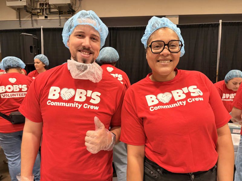 Two volunteers wearing red "BOB'S Community Crew" t-shirts, blue hairnets, and gloves, smiling at a community event.