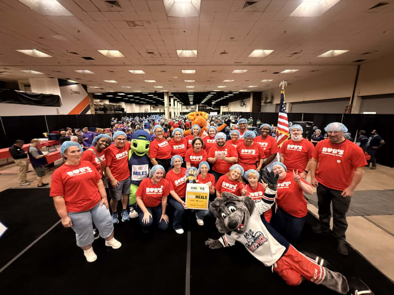Group of volunteers wearing red "Bob's Community Crew" shirts and blue hairnets posing indoors with mascots, holding a yellow box labeled "UNITE to Fight Hunger 20M Meals."