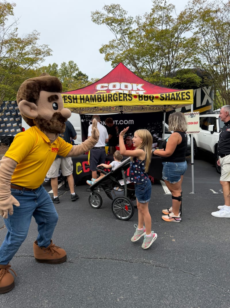 A person in a large mascot costume with brown hair, beard, and yellow shirt high-fives a young girl in a star-patterned tank top and denim shorts at a Cook Out tent offering fresh hamburgers, BBQ, and shakes.