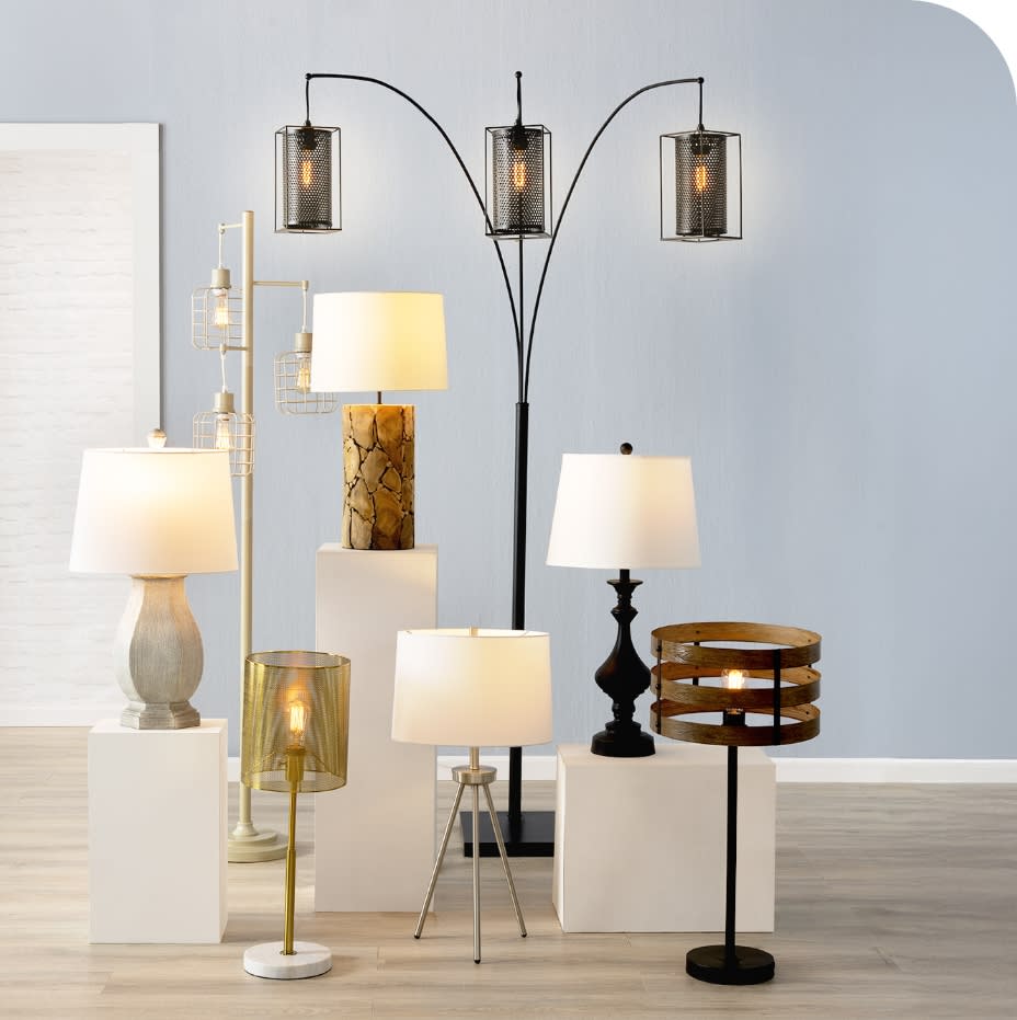 A collection of modern floor and table lamps with various designs, including metal mesh shades, wooden bases, and fabric lampshades, displayed on white pedestals against a light gray wall.