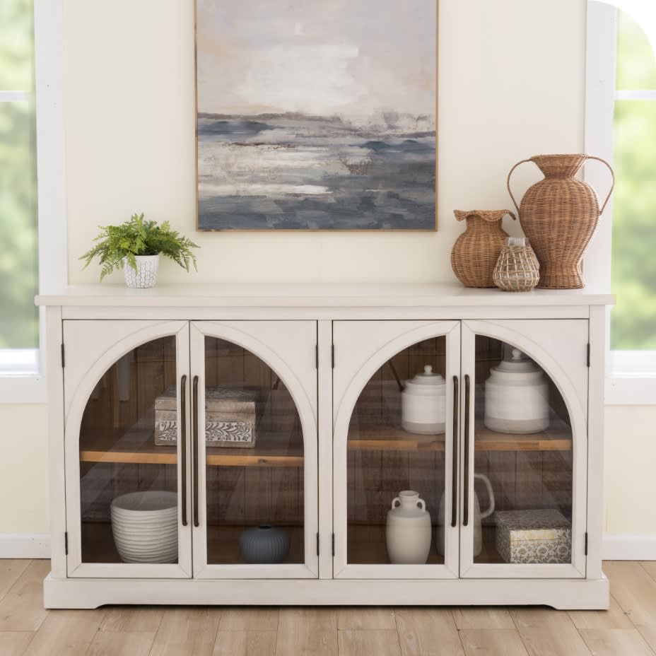 Black wooden cabinet with glass-paneled doors displaying ceramic vases and bowls, topped with a black and brass table lamp, decorative foliage, and a round wooden-framed mirror. Modern rustic home decor with natural wood backing and dark metal handles.