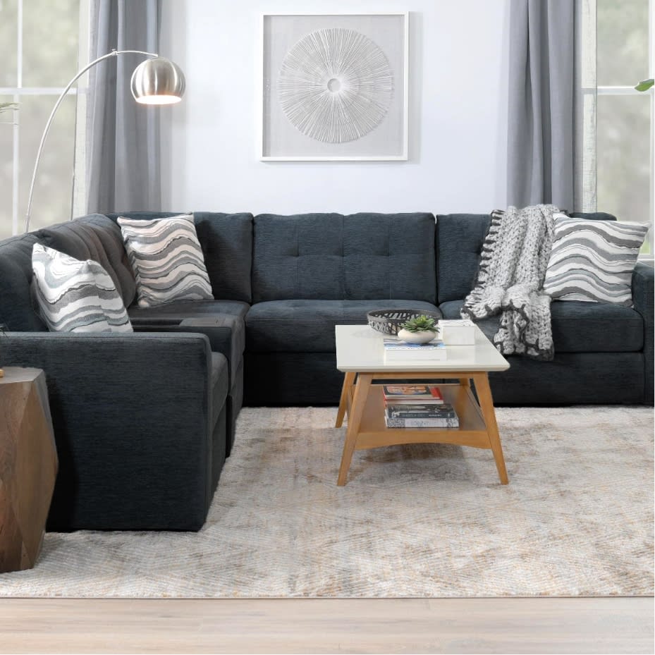 Modern living room with dark gray fabric sectional sofa, patterned throw pillows, a wooden coffee table with white top, and a textured area rug.