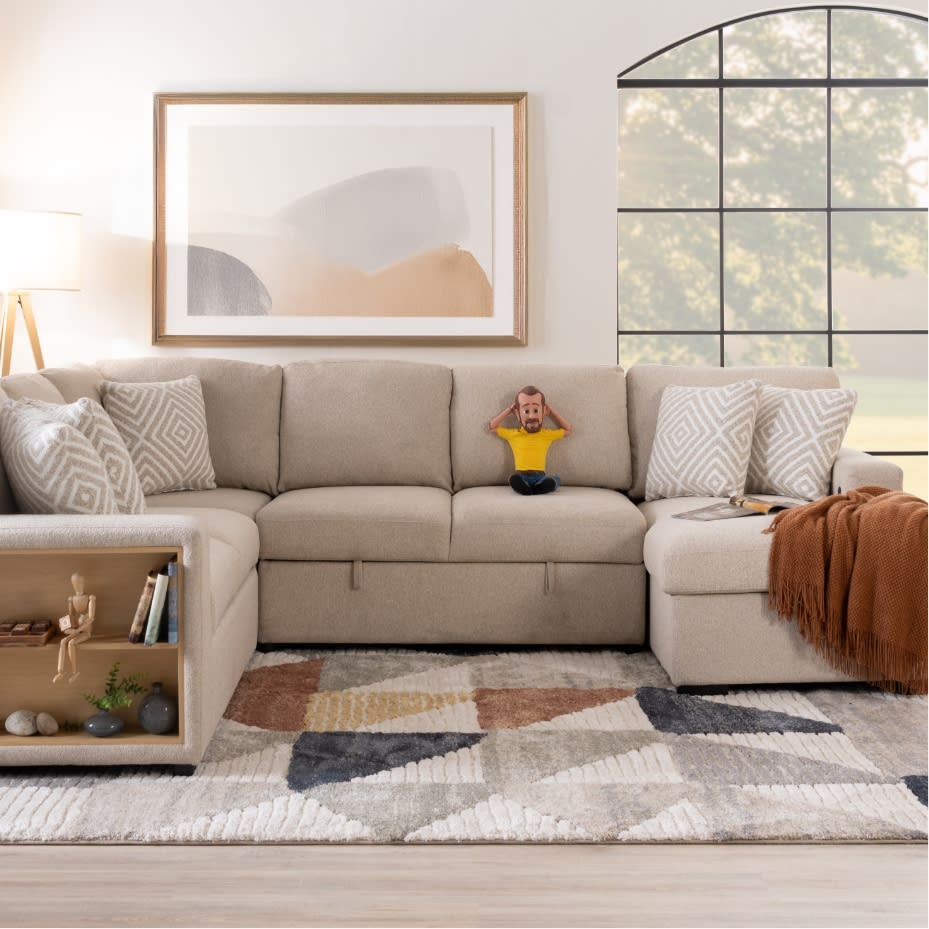 Modern beige fabric sectional sofa with multiple patterned throw pillows, a decorative wooden shelf, a framed abstract wall art, and a floor lamp.
