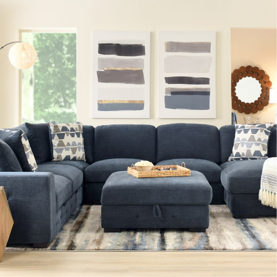 Modern living room with a large navy blue sectional sofa, patterned throw pillows, a matching ottoman, abstract wall art, and a woven tray with snacks.